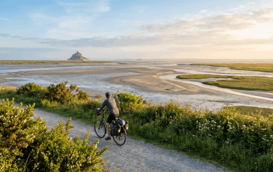Cycliste longeant la piste Vélomaritime avec vue sur le Mont-Saint-Michel au printemps, lumière dorée sur la baie bretonne
